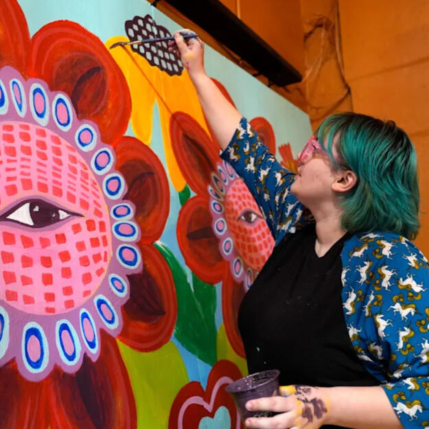 A blue haired person has their arm raised with a paintbrush about to touch the canvas. They are painting a figure with a bright red flower for a face. In the other hand, they hold a cup of dark purple paint.