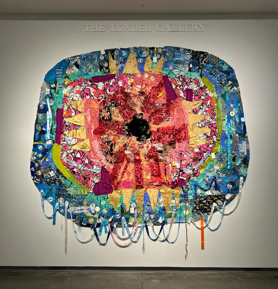 Bird Mother's Eye - Portal for MFA Thesis Show - Textile and Reclaimed Material Collage - 13ft x 13ft - 2025 A circular tapestry hangs on a whtie gallery wall. The tapestry is very colorful, covered in scraps of fabric that are red, pink, blue, green, white, and yellow. Ribbons hang down from the tapestry. At the center is a black pupil, making the tapestry look
