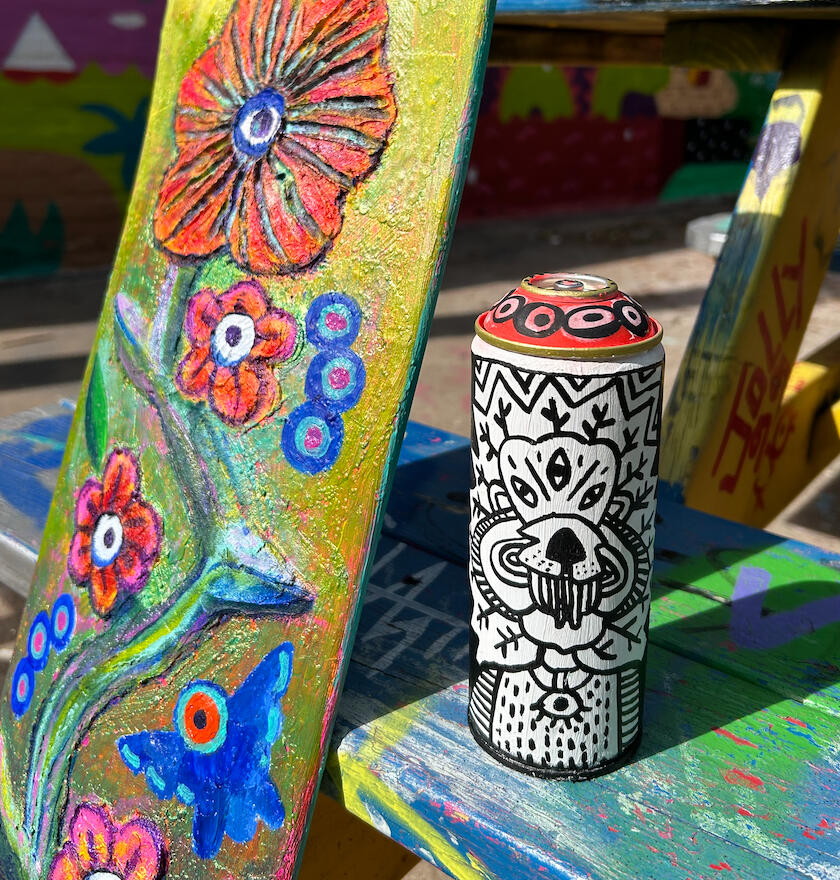 Image shows colorful skateboard with flowers and birds painted on it. A spraypaint can sits on a picnic bench with black and white drawings on it.